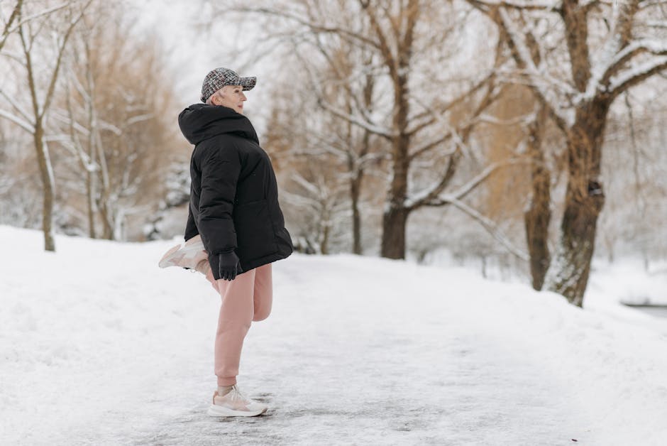 A woman dressed in a black winter jacket, beige sweatpants, and white sneakers is standing outdoors on a snow-covered path. She is wearing a checkered cap and black gloves, with her hands relaxed at her sides, gazing towards the distance. The background features snow-laden trees with bare branches, and the scene is illuminated by natural daylight, suggesting cold weather. The snowy environment and her attire indicate winter conditions suitable for outdoor activities. This image does not directly depict house removals or moving services but could relate to the winter timing of home relocation or packing and moving processes under adverse weather conditions, as managed by companies like Man With a Van Paddington.