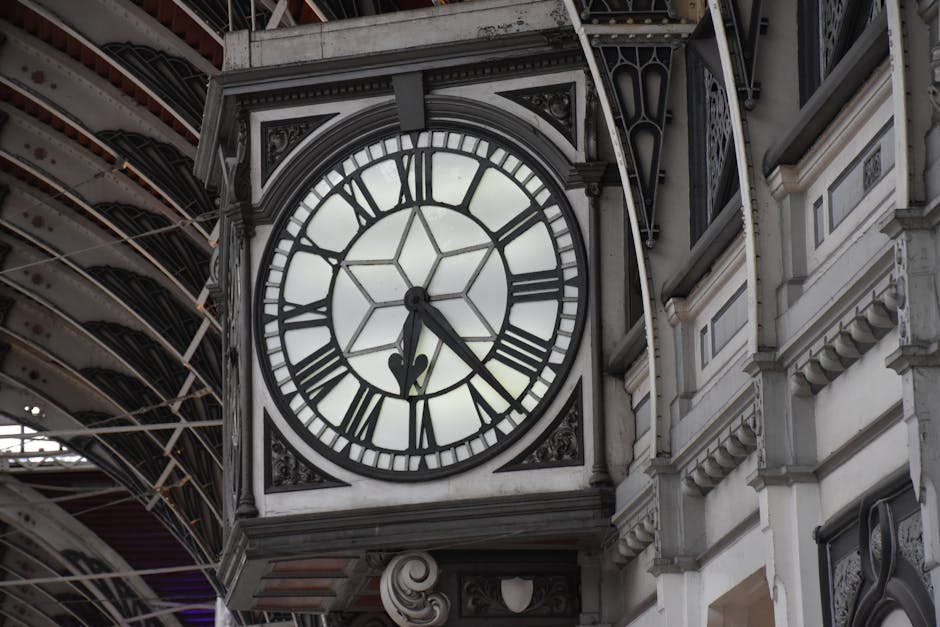 Close-up of a large, vintage clock mounted on the exterior of a building in Paddington, with Roman numerals and black clock hands showing the time as approximately 4:23. The clock face is surrounded by ornate, decorative metalwork and set against a backdrop of architectural details in white stone. Above the clock, there is a glimpse of the curved, iron-framed roof structure of Paddington train station, with visible metal supports and glass panels that allow natural light to illuminate the history-rich structure. The scene captures the historic railway station’s blend of classic design elements and modern infrastructure, illustrating the environment where [COMPANY_NAME], such as Man With a Van Paddington, may facilitate home relocation and furniture transport, including packing, loading, and moving logistics within this setting.