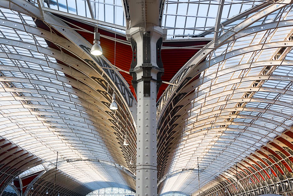 A detailed view of an indoor architectural structure focusing on a central metal column with rivets, supporting a complex roof framework made of curved metal and glass panels. The roof panels are transparent, allowing natural light to illuminate the space, with red accents visible in the ceiling area. Hanging lights are attached to the ceiling, and the structure appears to be part of a large transit station or public building. This setting is consistent with a busy urban environment where house removals and furniture transport services by Man With a Van Paddington would efficiently operate, facilitating packing and loading of goods for home relocation. The image emphasizes the industrial design and architectural detail relevant to moving logistics and transport processes.