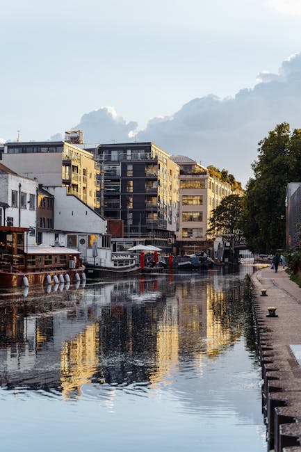 A canal scene featuring a row of modern multi-storey residential and commercial buildings with large windows and balconies, situated along the water's edge during daytime. Several boats, including a white passenger boat and smaller vessels, are moored along the canal. The water reflects the buildings and a partly cloudy sky with some sunlight illuminating the scene. On the right side, a paved pathway runs parallel to the canal, bordered by green trees and a low stone wall, with a few pedestrians walking. In the foreground, a small loader or trolley is visible near the edge of the water, possibly used for loading or unloading items for home relocation or furniture transport. This setting relates to house removals or moving services as part of a relocation process, with visible equipment and environment supporting loading, packing, and transport activities as seen in professional services such as Man With a Van Paddington.