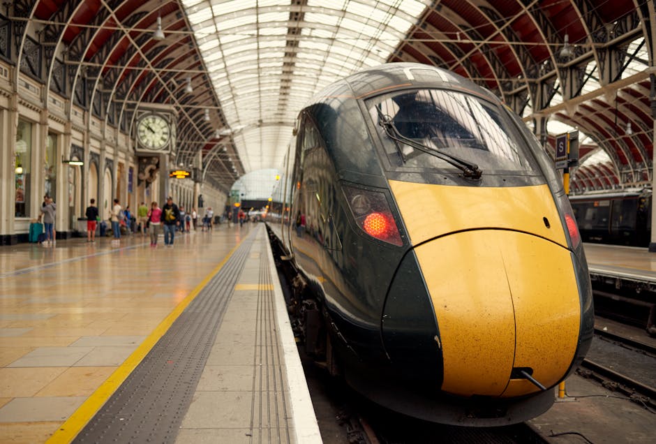Inside a historic train station with a high, arched glass roof and ornate architectural details, a modern high-speed train with a sleek black and yellow exterior is stopped at the platform. The train's front is rounded, with visible windshield wipers and illuminated red tail lights. The platform is lined with tactile paving and a yellow safety line along the edge. Several passengers are waiting on the platform, some standing near the train while others are seated on benches or walking. Baggage and parcels are visible in the foreground, some packed in cardboard boxes and plastic-wrapped items, indicating the process of packing and preparing for a move. A large clock hangs on the station wall, and additional trains or carriages can be seen further down the platform. The scene captures the busy environment of a major rail hub, with a focus on the logistics involved in travel and transportation, naturally aligning with house removals and moving services offered by Man With a Van Paddington.
