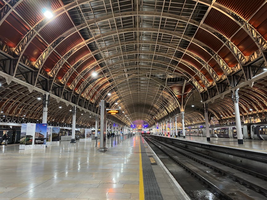 Interior view of a spacious train station with a high, arched ceiling made of metal and glass panels, illuminated by bright ceiling lights. The platform is clean and mostly empty, with a few passengers walking along the edge and some standing near the informational displays. On the right side, several modern train carriages are stationary, with closed doors ready for boarding. The station features multiple informational signs, including electronic departure boards, and promotional posters displayed on the walls. The smooth, polished floor and the yellow safety line along the platform indicate a well-maintained transport hub. This environment suggests an emphasis on efficient travel logistics, similar to those managed by Man With a Van Paddington for home relocation and furniture transport services, where effective moving operations and careful handling of items are essential.