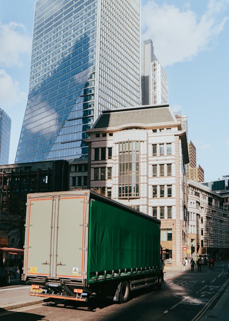 A large green shipping container truck is parked on a city street in front of tall modern office buildings with glass facades and older stone structures. The truck is positioned near the curb, ready for loading or unloading, with its rear doors closed. The surrounding environment includes pedestrians walking on the sidewalk and other vehicles on the road, typical of an urban setting in central London. The scene depicts a home relocation process involving furniture or household items being transported as part of a professional removals service, with natural daylight highlighting the buildings and vehicle. Man With a Van Paddington specializes in residential and business removals, supporting clients with furniture transport and packing logistics within the city.