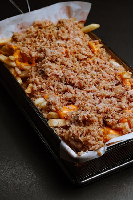 A close-up view of a food container placed on a black surface, filled with a loaded tray of loaded fries topped with shredded cheese, crispy fried onion strings, and crumbled bacon bits. The fries are covered with orange cheese sauce and garnished with seasoning. The container appears to be a takeaway tray lined with parchment paper, and the background includes a dark surface with no additional objects visible. This image focuses on the presentation of the loaded fries, evoking a snack or fast-food appearance, with no elements related to house removals, packing, or transport services. The image highlights the texture of the crispy toppings and the melted cheese, suitable for food-related content but does not align with relocation or moving themes.