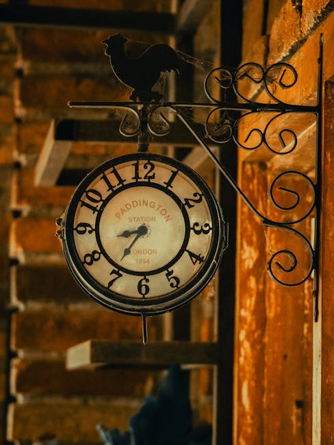 A vintage-style round clock with a white face, black numerals, and black hands, is mounted on a decorative wrought iron bracket affixed to a brick wall. The clock features the words 'Paddington Station' and 'London 1854' on its face. Above the clock, there is a small black silhouette of a dog or fox, also attached to the wrought iron bracket, which has elegant scrollwork. The setting appears to be indoors, possibly in a station or a building with exposed brickwork and wooden beams in the background, illuminated with warm lighting. This clock installation relates to house removals or moving services by emphasizing the importance of timing and coordination during a relocation, as represented by the vintage clock and decorative elements.