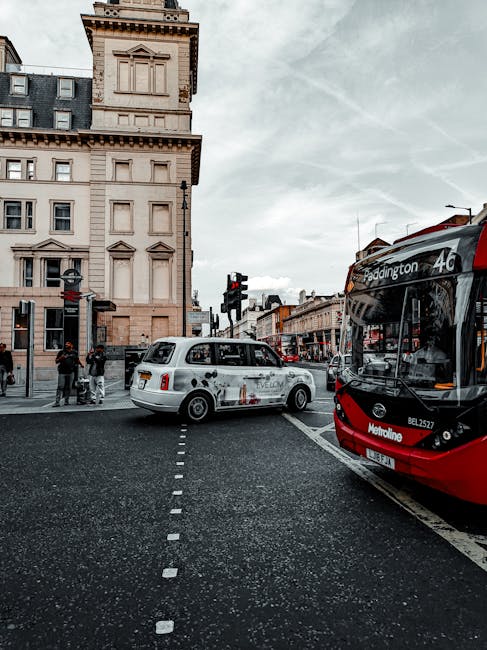 A street scene in Paddington featuring a red and black double-decker bus with route signage indicating it serves Paddington. To the left, a white luxury van with company branding is parked near a historic, multi-storey building with ornate architectural details. Several pedestrians are waiting at a nearby crosswalk; some are standing on the pavement, while others are preparing to cross. The road has marked lanes and is slightly wet, suggesting recent rain. Traffic lights are visible, controlling vehicle and pedestrian movement. The overall environment reflects an urban setting with a mix of transportation modes, emphasizing busy city life and logistical elements associated with house removals and packing services. The scene highlights the transportation aspect of home relocation, involving vehicle loading and passenger movement, consistent with services offered by Man With a Van Paddington.