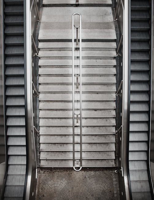 A view from above of a stairwell with a metal handrail running down the center, flanked by two sets of stairs with grey concrete steps. The steps have textured surfaces for grip, and the sides are enclosed by metallic railings. The staircase is situated within an indoor environment, with illuminated lighting casting shadows, and the surrounding area appears to be part of a residential or commercial building lobby. The image captures the process of moving or transporting furniture and boxes, which are not directly visible in this shot, but the setting suggests an ongoing or planned home relocation. The stairwell's utilitarian design, along with the visible materials, highlights the typical corridor used in internal building moves, as managed by professional removal services such as Man With a Van Paddington, especially when navigating tight stair access during furniture transport and packing and moving activities.