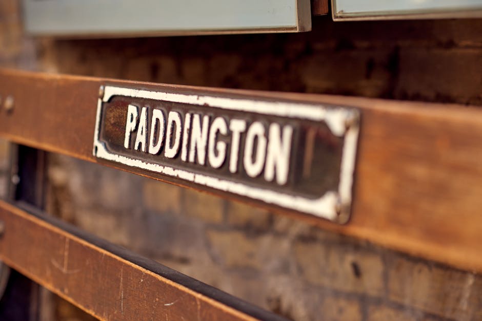 Close-up of a vintage metal shipping crate with a rusted finish, mounted on a wooden pallet. The crate features a black and white label with the word 'PADDINGTON' in bold, uppercase letters, slightly worn and weathered. The wooden pallet shows signs of use, with visible grain and aged surface, and is situated indoors near a brick wall with warm lighting. The scene suggests a setting related to home removal logistics, packing, or transporting goods during a move, with the crate possibly used for storing or shipping items as part of furniture transport or packing and moving services offered by Man With a Van Paddington.