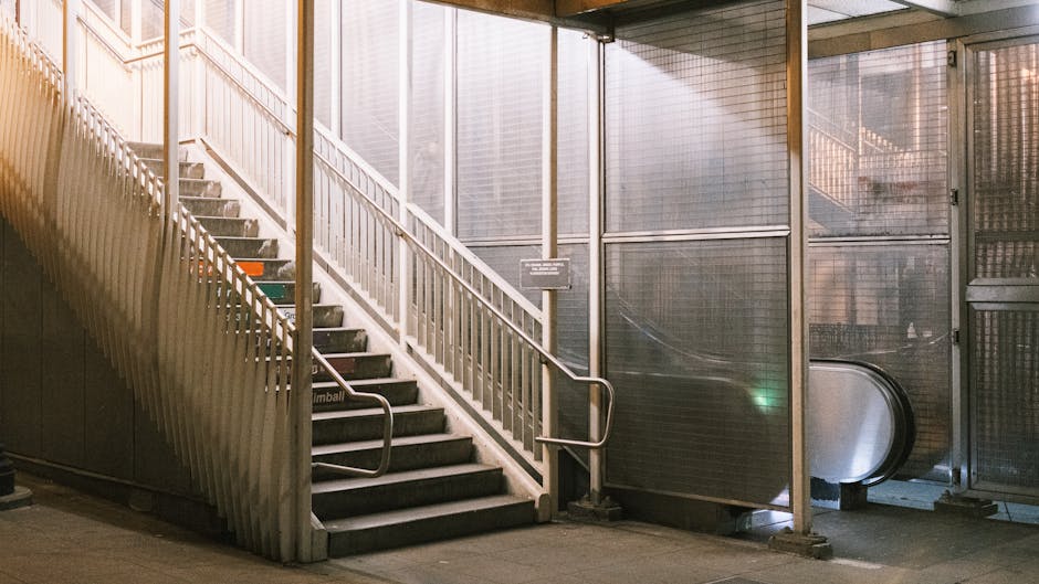 A view from above of a stairwell with a metal handrail running down the center, flanked by two sets of stairs with grey concrete steps. The steps have textured surfaces for grip, and the sides are enclosed by metallic railings. The staircase is situated within an indoor environment, with illuminated lighting casting shadows, and the surrounding area appears to be part of a residential or commercial building lobby. The image captures the process of moving or transporting furniture and boxes, which are not directly visible in this shot, but the setting suggests an ongoing or planned home relocation. The stairwell's utilitarian design, along with the visible materials, highlights the typical corridor used in internal building moves, as managed by professional removal services such as Man With a Van Paddington, especially when navigating tight stair access during furniture transport and packing and moving activities.