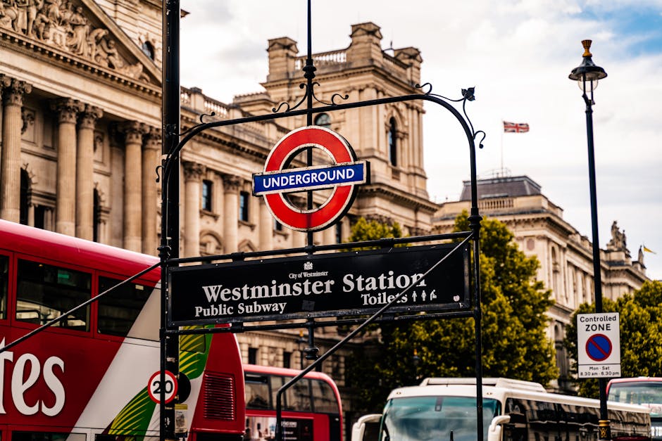 A street scene outside Westminster Station in London, featuring a red double-decker bus partially visible on the left, parked on the pavement. Above, there is a black hanging sign with the London Underground roundel logo and a smaller sign reading 'Westminster Station', indicating a public subway entrance. The background showcases large, neoclassical-style buildings with detailed stone facades, decorative columns, and a Union Jack flag flying atop one of the structures. The scene is illuminated by daylight, with clear blue sky and some cloud coverage. To the right, a traditional black lamppost and various street signs, including a controlled zone sign, are visible. This setting relates to urban transportation and city logistics, with the presence of the underground signage subtly aligning with house removals and relocation services such as those offered by Man With a Van Paddington, supporting efficient furniture transport and loading processes.