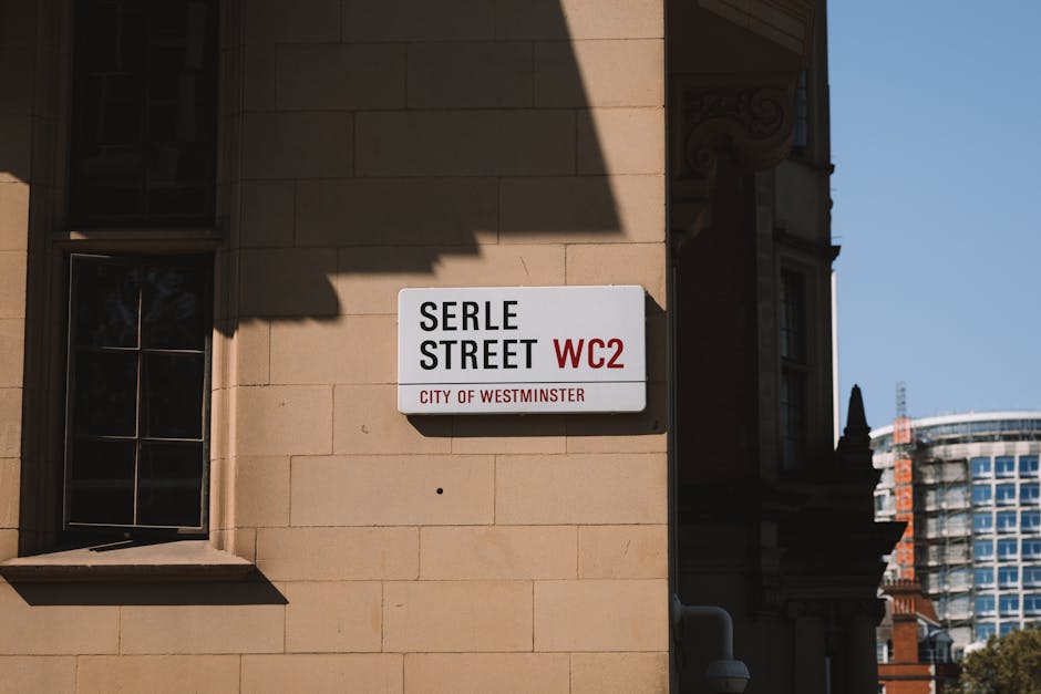 A street sign mounted on a beige stone building corner displaying 'Serle Street WC2, City of Westminster'. The sign is white with black and red text, indicating the street name and postal code. To the left, a small window with black metal bars and a window sill cast shadows due to sunlight. Adjacent to the sign, part of an ornate building facade with decorative architectural details is visible, with another building with large glass windows and a modern structure seen in the background. The scene captures an urban environment relevant to house removals and local moving services, illustrating a typical London street setting associated with property relocations. The lighting suggests a clear day, and the overall view reflects an area where furniture transport and packing processes could be underway as part of a home relocation or removal service.