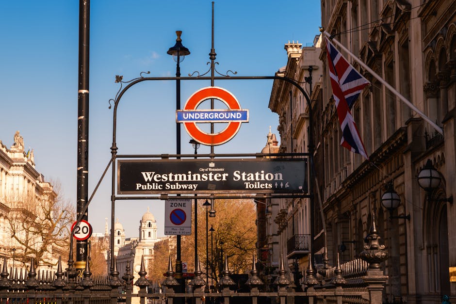 A street scene outside Westminster Station in London, featuring a red double-decker bus partially visible on the left, parked on the pavement. Above, there is a black hanging sign with the London Underground roundel logo and a smaller sign reading 'Westminster Station', indicating a public subway entrance. The background showcases large, neoclassical-style buildings with detailed stone facades, decorative columns, and a Union Jack flag flying atop one of the structures. The scene is illuminated by daylight, with clear blue sky and some cloud coverage. To the right, a traditional black lamppost and various street signs, including a controlled zone sign, are visible. This setting relates to urban transportation and city logistics, with the presence of the underground signage subtly aligning with house removals and relocation services such as those offered by Man With a Van Paddington, supporting efficient furniture transport and loading processes.